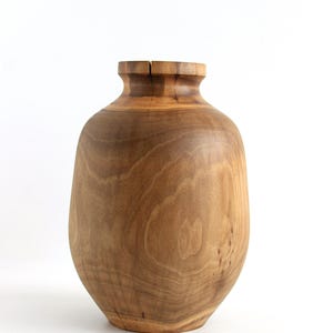 May include: A handcrafted wooden vase with a natural finish. The vase has a rounded body and a narrower neck, with visible wood grain patterns. The vase is approximately 10 inches tall and is suitable for home decor.