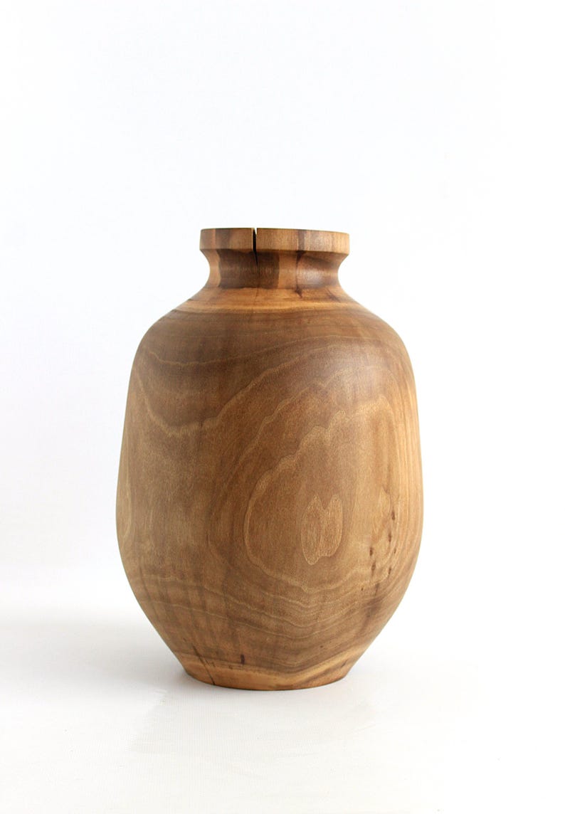 May include: A handcrafted wooden vase with a natural finish. The vase has a rounded body and a narrower neck, with visible wood grain patterns. The vase is approximately 10 inches tall and is suitable for home decor.
