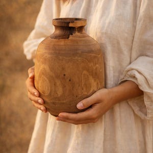 May include: A handcrafted wooden vase with a rounded body and a narrow neck. The vase is made of light brown wood with visible grain patterns. The vase is held in front of a person wearing a cream-colored dress.