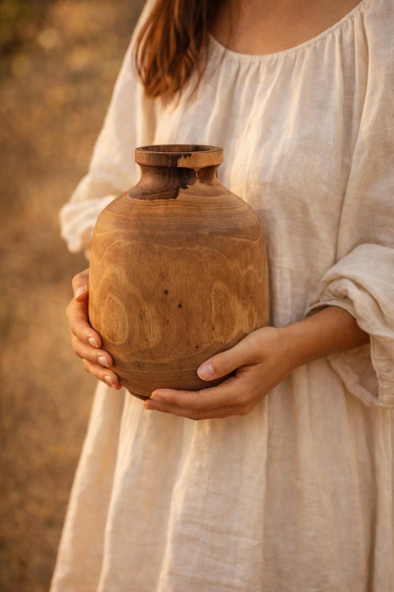 May include: A handcrafted wooden vase with a rounded body and a narrow neck. The vase is made of light brown wood with visible grain patterns. The vase is held in front of a person wearing a cream-colored dress.