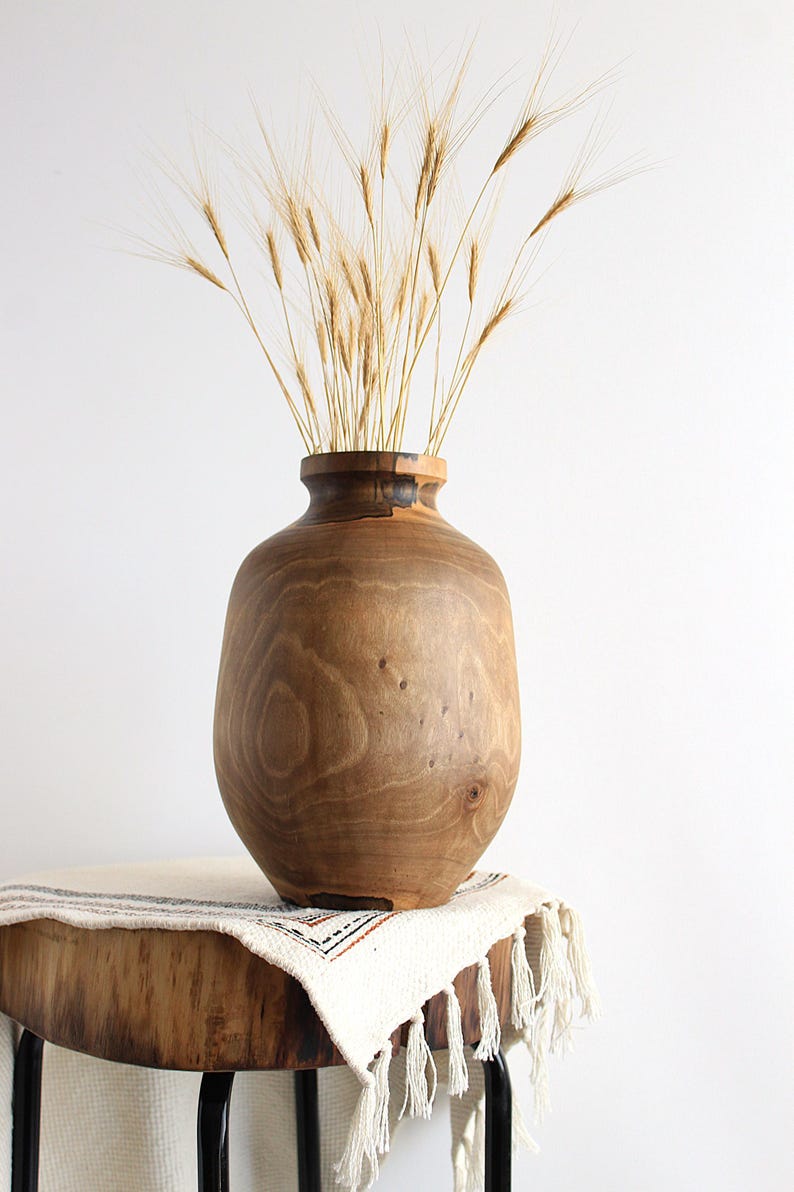 May include: A large, handcrafted wooden vase with a wide, rounded body and a narrow neck. The vase is filled with dried wheat stalks, creating a natural, rustic aesthetic. The vase is displayed on a wooden table with a fringed cloth.