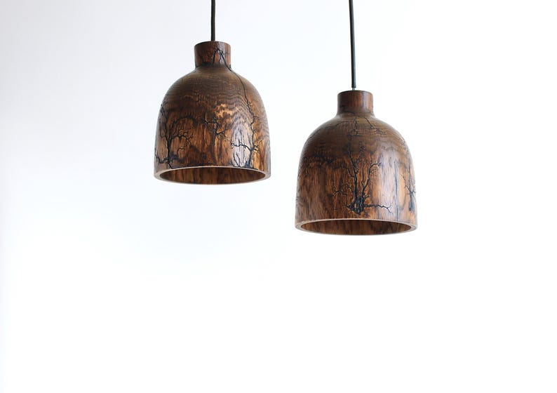 Peut inclure: Deux suspensions en forme de d&ocirc;me fabriqu&eacute;es en bois brun fonc&eacute;, chacune pr&eacute;sentant des motifs fractals noirs complexes sur la surface. Les luminaires sont suspendus par de fins cordons noirs, mettant en valeur le grain naturel du bois.