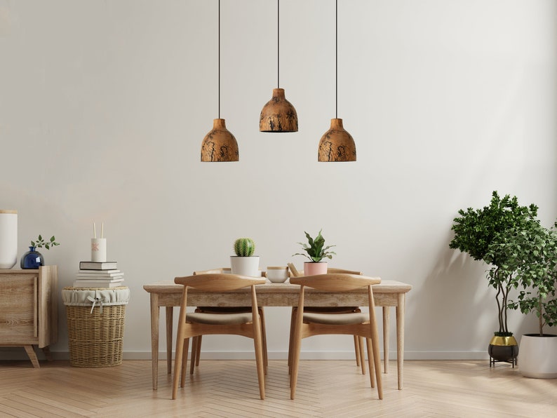 Peut inclure: Trois suspensions en forme de d&ocirc;me avec une finition brune textur&eacute;e sont suspendues au-dessus d'une table &agrave; manger en bois clair. La table est accompagn&eacute;e de deux chaises en bois avec dossiers incurv&eacute;s et coussins beiges, avec des plantes.