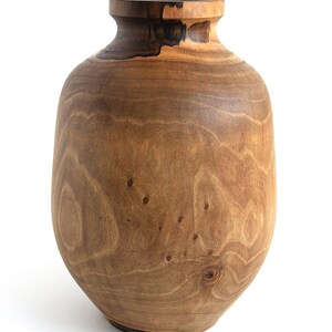 May include: A handcrafted wooden vase with a rounded body and a narrow neck. The vase is made of light brown wood with darker brown accents and visible wood grain. The vase is a decorative item.