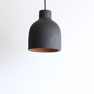 Peut inclure: Une suspension en forme de cloche avec un ext&eacute;rieur noir mat pr&eacute;sentant une texture de grain de bois vertical. L'int&eacute;rieur de l'abat-jour r&eacute;v&egrave;le une finition en bois brun clair naturel. La lampe est suspendue par un fin cordon noir sur fond blanc.