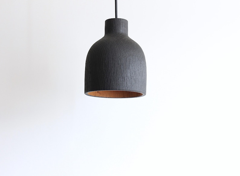 Peut inclure: Une suspension en forme de cloche avec un ext&eacute;rieur noir mat pr&eacute;sentant une texture de grain de bois vertical. L'int&eacute;rieur de l'abat-jour r&eacute;v&egrave;le une finition en bois brun clair naturel. La lampe est suspendue par un fin cordon noir sur fond blanc.