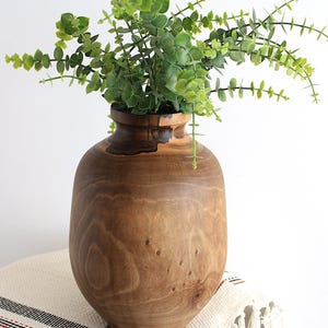 May include: A large, wooden vase with a wide, rounded body and a narrow neck, filled with green leafy branches. The vase sits on a small, round table covered with a cream-colored cloth with black and brown stripes.