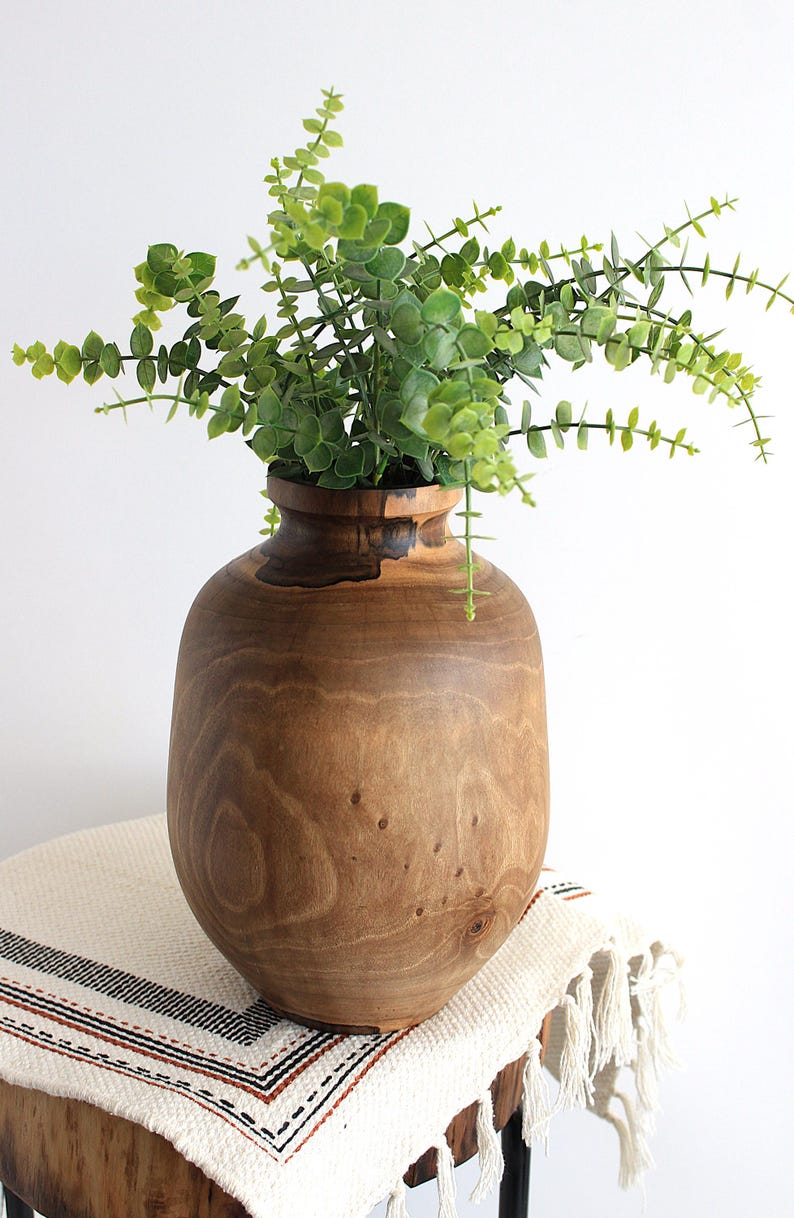May include: A large, wooden vase with a wide, rounded body and a narrow neck, filled with green leafy branches. The vase sits on a small, round table covered with a cream-colored cloth with black and brown stripes.