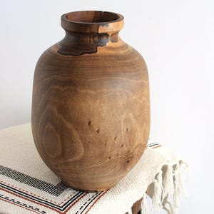 May include: A large, handcrafted wooden vase with a wide, rounded body and a narrow neck. The vase is made of light brown wood with darker brown accents and visible wood grain. It sits on a small, round wooden table covered with a cream-colored textile.
