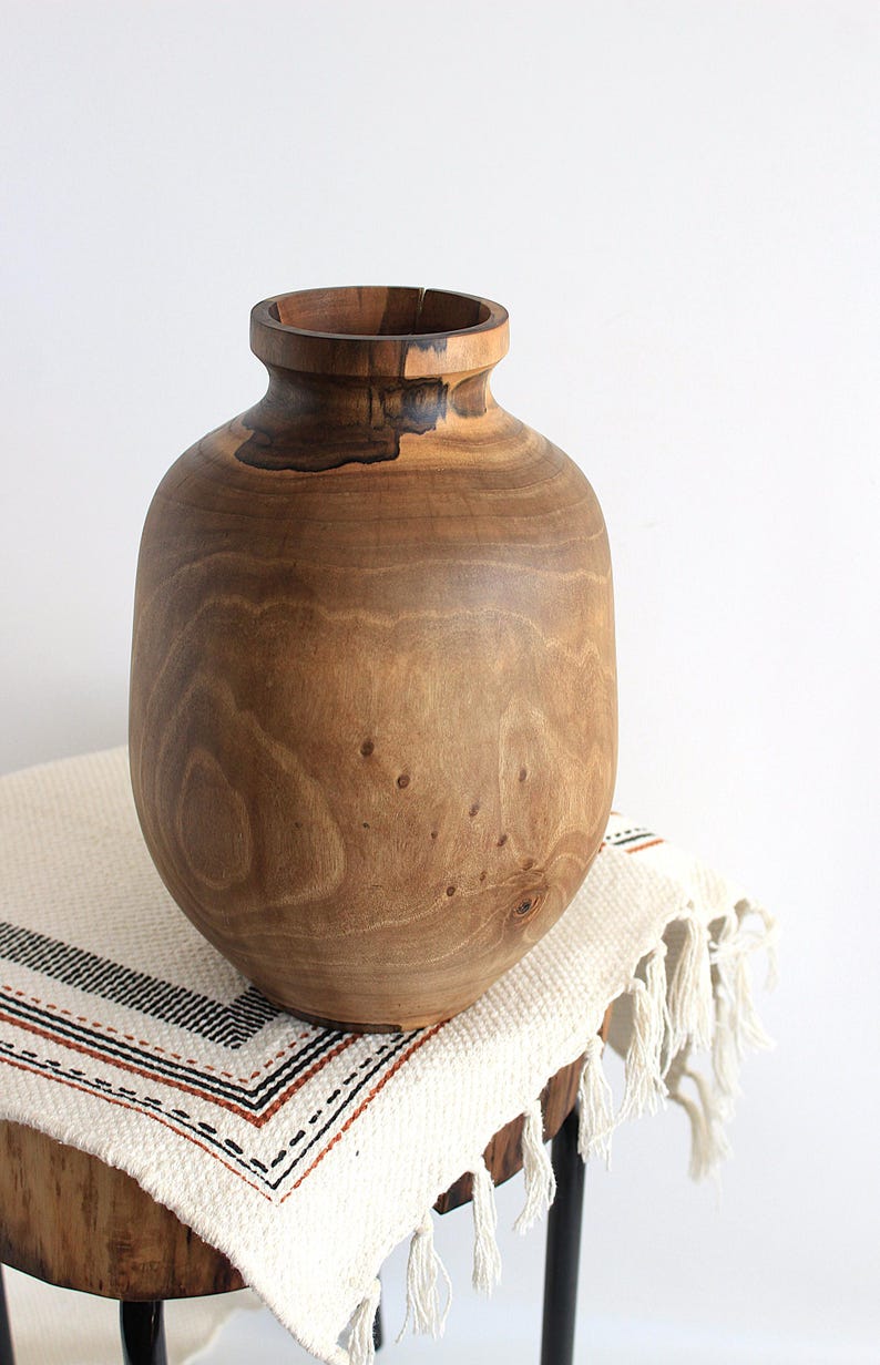 May include: A large, handcrafted wooden vase with a wide, rounded body and a narrow neck. The vase is made of light brown wood with darker brown accents and visible wood grain. It sits on a small, round wooden table covered with a cream-colored textile.