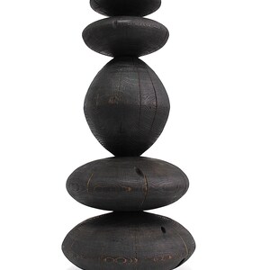 May include: A stacked sculpture composed of six dark, oval-shaped wooden pieces. The pieces are arranged vertically, creating a balanced, abstract form. The wood grain is visible, adding texture to the dark, almost black, surface. The sculpture is set against a white background.