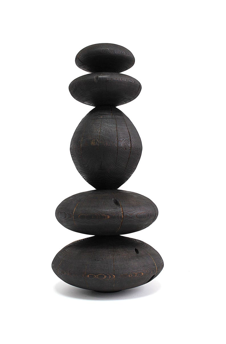May include: A stacked sculpture composed of six dark, oval-shaped wooden pieces. The pieces are arranged vertically, creating a balanced, abstract form. The wood grain is visible, adding texture to the dark, almost black, surface. The sculpture is set against a white background.