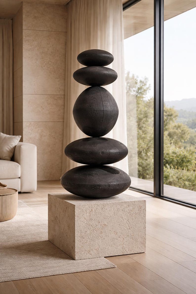 May include: A modern sculpture featuring a stack of five dark, oval-shaped stones atop a square, light-colored pedestal. The stones vary in size and are arranged to create a balanced, artistic display. The sculpture is a decorative element for interior spaces.