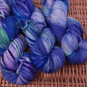 May include: A collection of hand-dyed yarn skeins in shades of blue, purple, and green. The yarn is arranged on a woven wicker surface, showcasing the vibrant colors and textures of the fiber. Ideal for knitting and crochet projects.