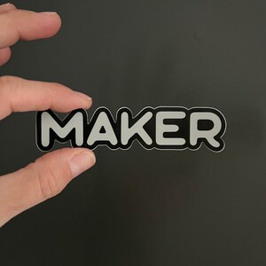 May include: A black and white sticker with the word "MAKER" in white letters with a black outline.