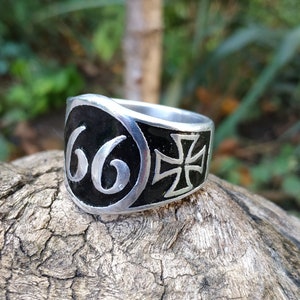 May include: A silver ring with a black enamel design. The ring features the number "66" on one side and a black cross on the other side.