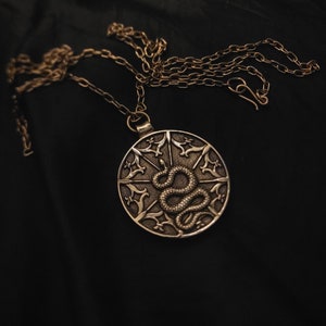 Puede incluir: Un collar de plata con un colgante redondo que presenta un diseño detallado de una serpiente enroscada alrededor de una estrella de explosión. El colgante está en una cadena con un diseño delicado e intrincado.