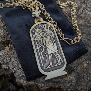 Op de afbeelding: Een goudkleurige hangerketting met een Egyptisch hiërogliefenontwerp. De rechthoekige hanger heeft een gegraveerde figuur en piramide. De ketting en sluiting zijn ook goudkleurig. De hanger ligt op een zwart fluwelen zakje, tegen een houten achtergrond.