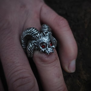 Op de afbeelding: Een zilveren ring met een schedelontwerp en rode ogen. De schedel heeft hoorns aan beide kanten. De ring wordt aan een vinger gedragen.