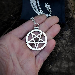 Puede incluir: Un collar de cadena de plata con un colgante de pentagrama. El colgante es una estrella de cinco puntas con un círculo alrededor.