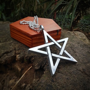 Può includere: Una collana con un ciondolo a forma di pentagramma argentato con una catena. Il ciondolo ha la forma di una stella a cinque punte con un design a nodo. La collana è appoggiata su una scatola di legno.