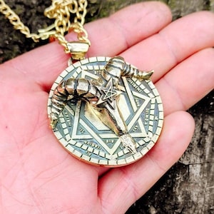 May include: A gold chain necklace with a pendant featuring a stylized ram skull with horns and a pentagram inside a circle with text around the edge.