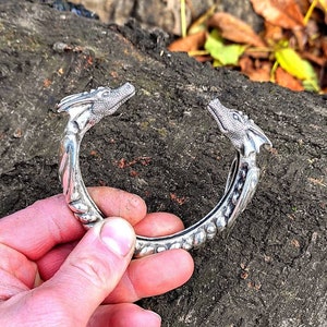 May include: A silver dragon bracelet featuring two dragon heads facing each other. The bracelet is crafted from thick, textured metal and boasts a detailed design.