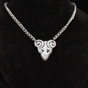 Puede incluir: Un collar de cadena de plata con un colgante de plata con forma de cabeza de carnero con cuernos rizados.