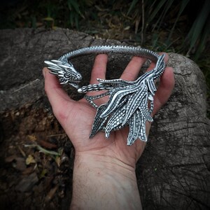 May include: Silver dragon necklace with a detailed design. The dragon's head is facing forward with its wings spread out behind it.