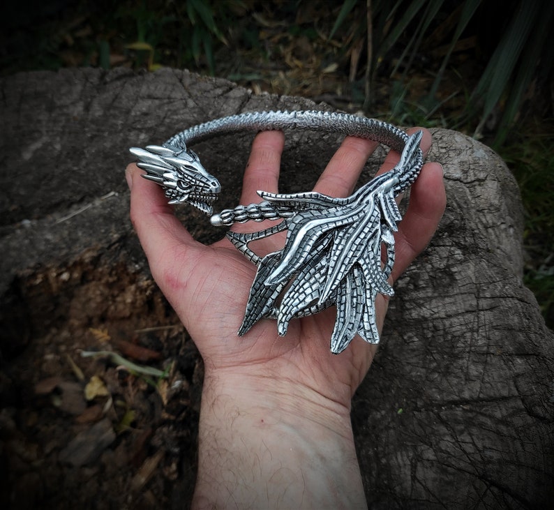May include: Silver dragon necklace with a detailed design. The dragon's head is facing forward with its wings spread out behind it.