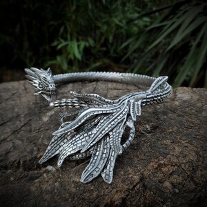May include: Silver dragon head necklace with a detailed design. The necklace is made of metal and has a dragon head with open mouth and sharp teeth. The dragon's wings are spread out and the necklace is open in the back.