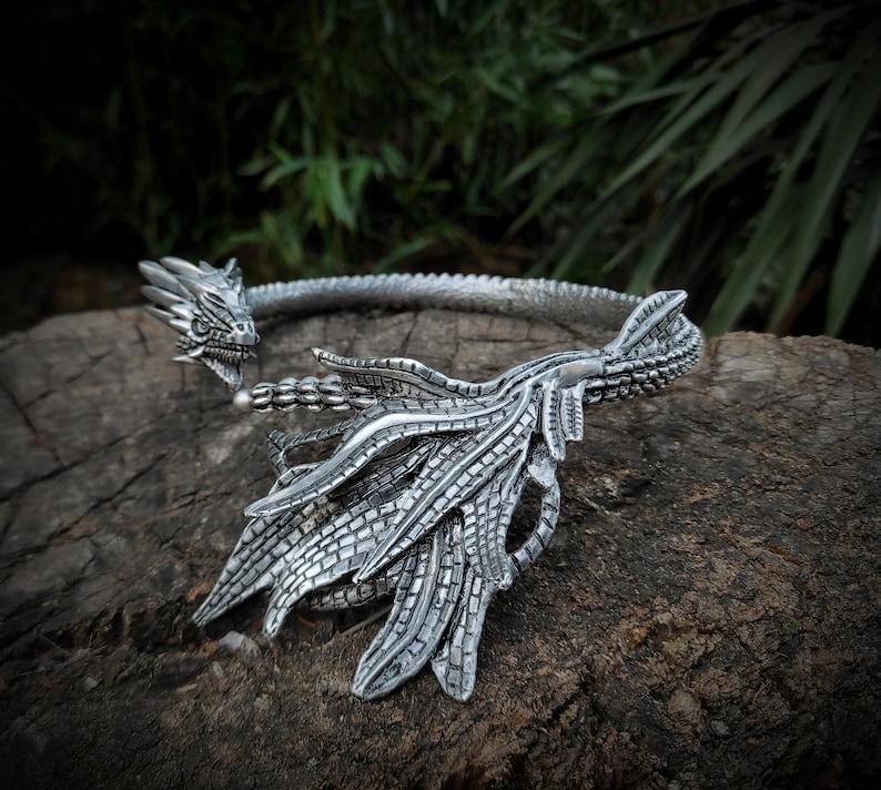 May include: Silver dragon head necklace with a detailed design. The necklace is made of metal and has a dragon head with open mouth and sharp teeth. The dragon's wings are spread out and the necklace is open in the back.