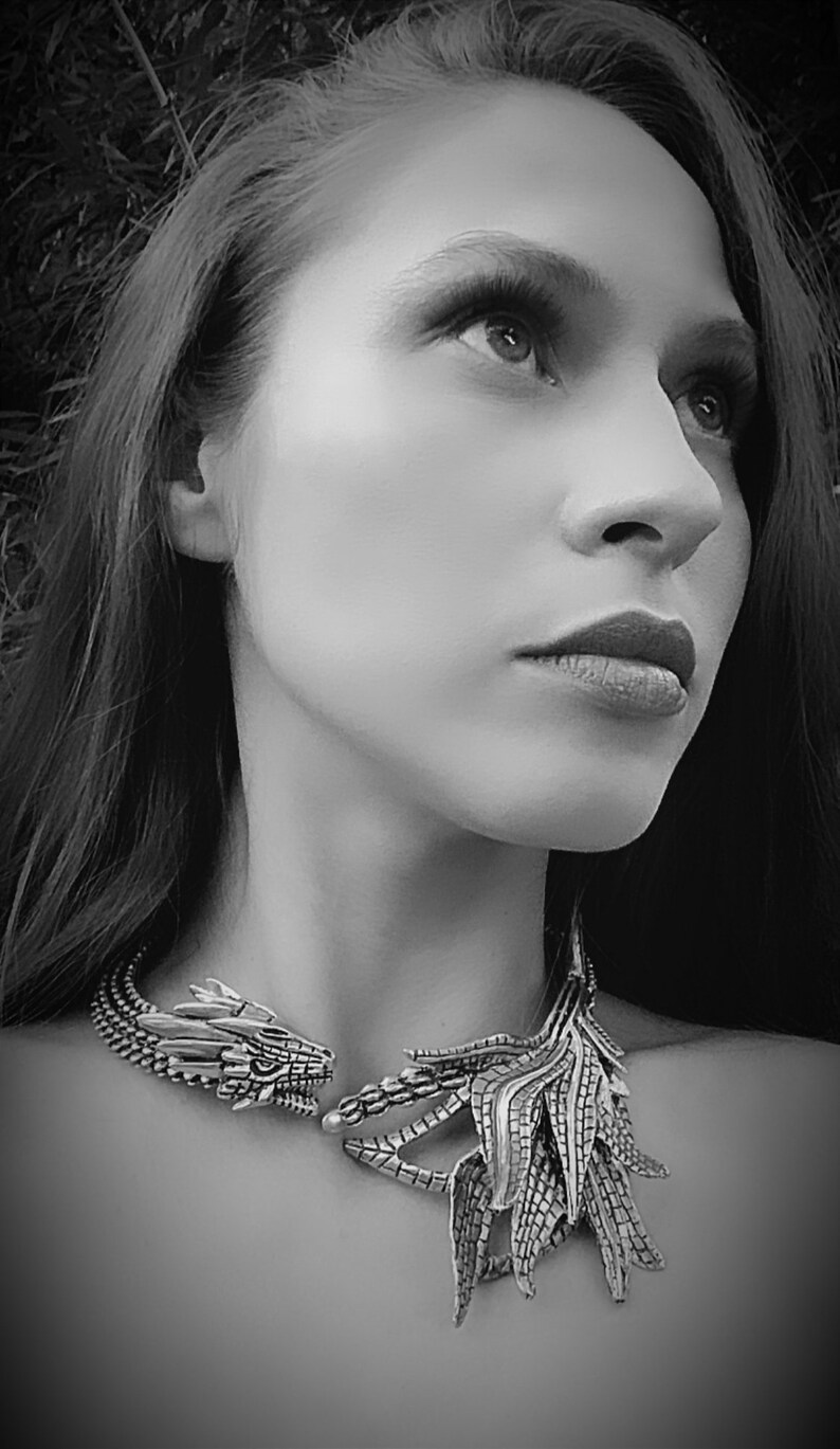 May include: A woman wearing a silver dragon necklace with a detailed design. The necklace features a dragon head on one side and a wing-like design on the other.