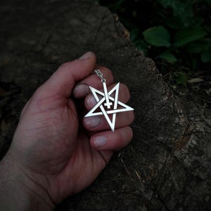 May include: A silver pendant necklace featuring a pentagram with an inverted cross inside. The pendant is held in a hand against a wooden background.