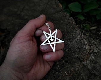 May include: A silver pendant necklace featuring a pentagram with an inverted cross inside. The pendant is held in a hand against a wooden background.