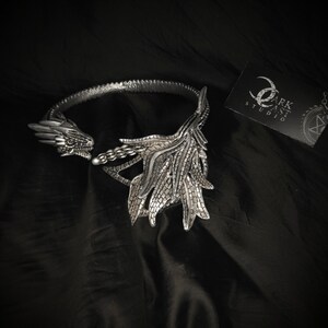 May include: Silver dragon necklace with a detailed design. The necklace is open in the back and has a dragon head on each side. The dragon's wings are spread out and have a scaly texture. The necklace is on a black fabric background.