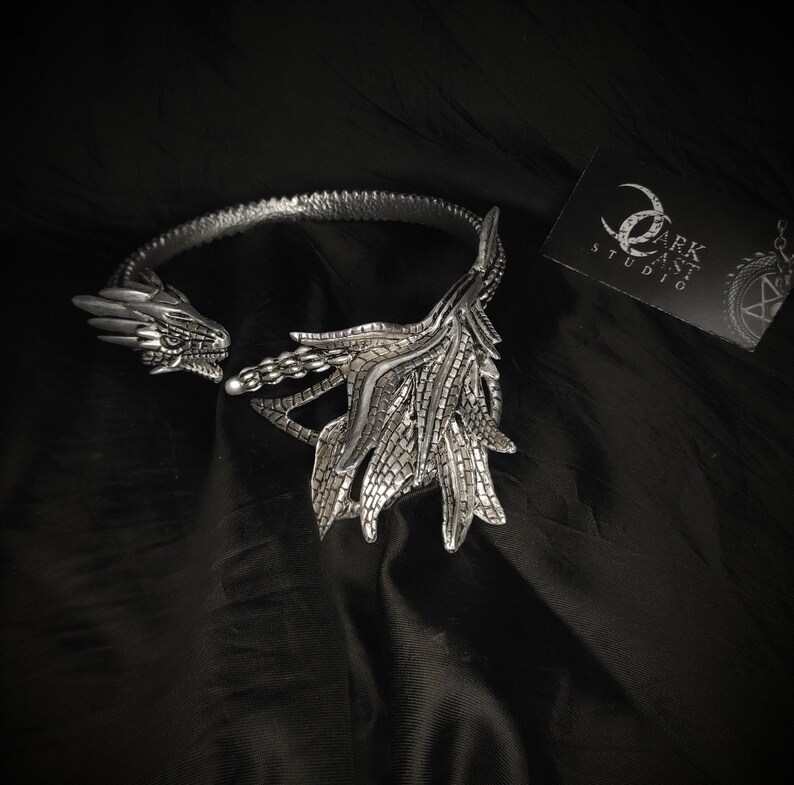 May include: Silver dragon necklace with a detailed design. The necklace is open in the back and has a dragon head on each side. The dragon's wings are spread out and have a scaly texture. The necklace is on a black fabric background.