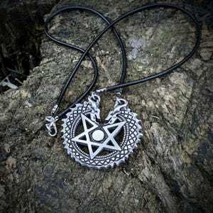 Puede incluir: Un colgante de metal plateado con dos cabezas de dragón enfrentadas que forman una figura circular, con una estrella de cinco puntas en el centro y un pequeño círculo en medio. El colgante está unido a un collar de cordón negro fino con cierres metálicos.