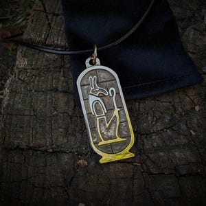 Puede incluir: Un colgante plateado y dorado con un diseño jeroglífico egipcio. El colgante es rectangular con bordes redondeados y cuelga de un cordón negro. El fondo es una superficie de madera oscura y texturizada.