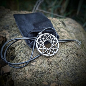 Può includere: Una collana con pendente color argento con un design a stella e un cordino nero. Il pendente circolare presenta un motivo a stella geometrica. Un sacchetto di velluto nero è dietro la collana, appoggiato su una superficie marrone strutturata.