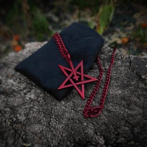 Op de afbeelding: Een rode metalen ketting met een pentagram hanger. De hanger heeft een kruis in de ster. De ketting ligt op een donker oppervlak.