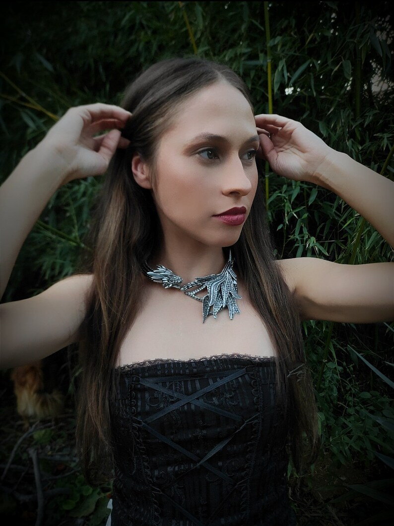 May include: A woman with long brown hair wearing a black corset top and a silver dragon necklace. The necklace has a detailed design with wings and a head.