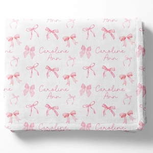 Könnte beinhalten: Eine weiße Babydecke mit einem Muster aus rosa Aquarell-Schleifen und dem Namen "Caroline Ann", der sich wiederholt.