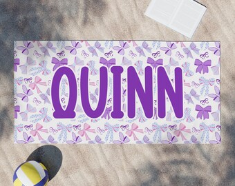 May include: A white beach towel with a purple bow pattern and the name "QUINN" printed in large purple letters.