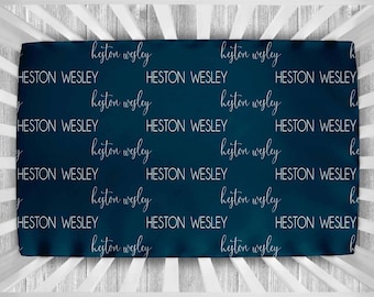 May include: A dark blue crib sheet with the name "Heston Wesley" repeated in white script throughout the fabric.