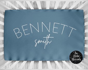 May include: A blue fitted crib sheet with the name "BENNETT" in large white capital letters and the name "smith" in a smaller white cursive font. The sheet has a logo in the bottom right corner that says "The Little Arrows".