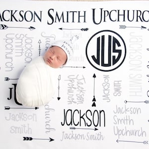 May include: A white baby blanket with black and gray text that reads "Jackson Smith Upchurch" and includes arrows and a monogram. A baby is lying on the blanket wearing a white hat with the name "Jackson" on it.
