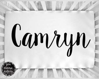 May include: White crib sheet with the name "Camryn" printed in black cursive font.