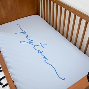 May include: A light blue cot sheet with the name "Peyton" written in blue cursive script. The sheet is on a wooden cot with a white wall in the background. The logo "The Little Arrows" is in the bottom right corner.