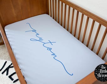 May include: A light blue crib sheet with the name "Peyton" written in blue cursive script. The sheet is on a wooden crib with a white wall in the background. The logo "The Little Arrows" is in the bottom right corner.